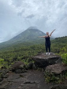 A young woman wearing athletic clothes posing on a rock in front of Arenal Volcano